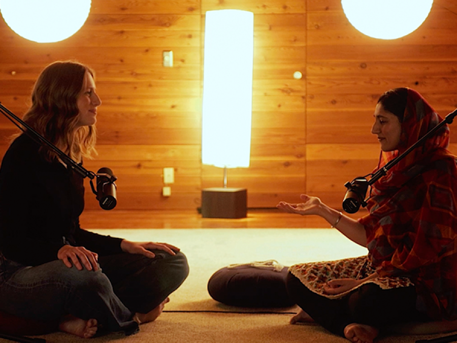 Two females sit on the floor cross legged speaking into microphones